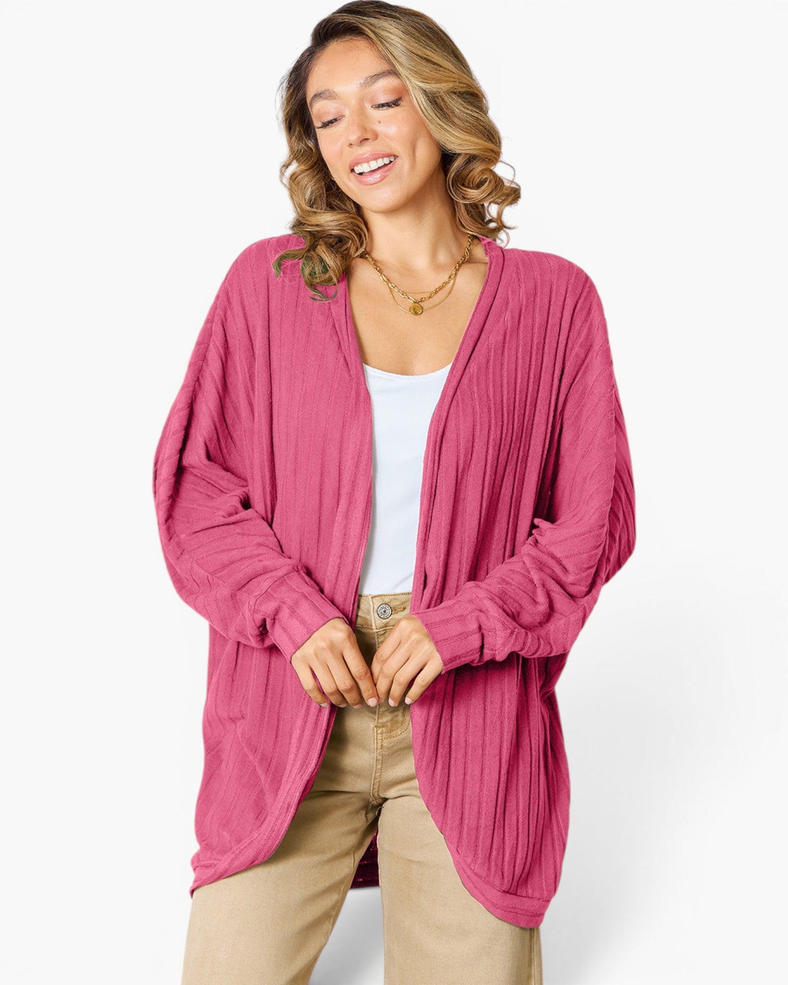A woman wearing a pink pleated cardigan over a white top and beige pants, standing against a light gray studio background.