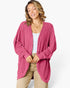 A woman wearing a pink pleated cardigan over a white top and beige pants, standing against a light gray studio background.