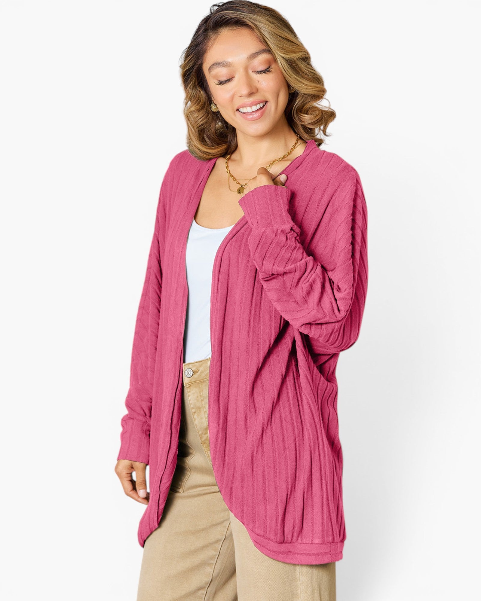 A woman wearing a pink long-sleeve open-front cardigan over a white tank top and beige pants, photographed against a clean white studio background.