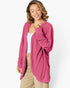 A woman wearing a pink long-sleeve open-front cardigan over a white tank top and beige pants, photographed against a clean white studio background.
