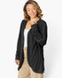 A woman wearing a black long-sleeve open-front cardigan with a textured vertical pleated pattern, styled over a white tank top and beige pants, against a plain white studio background.