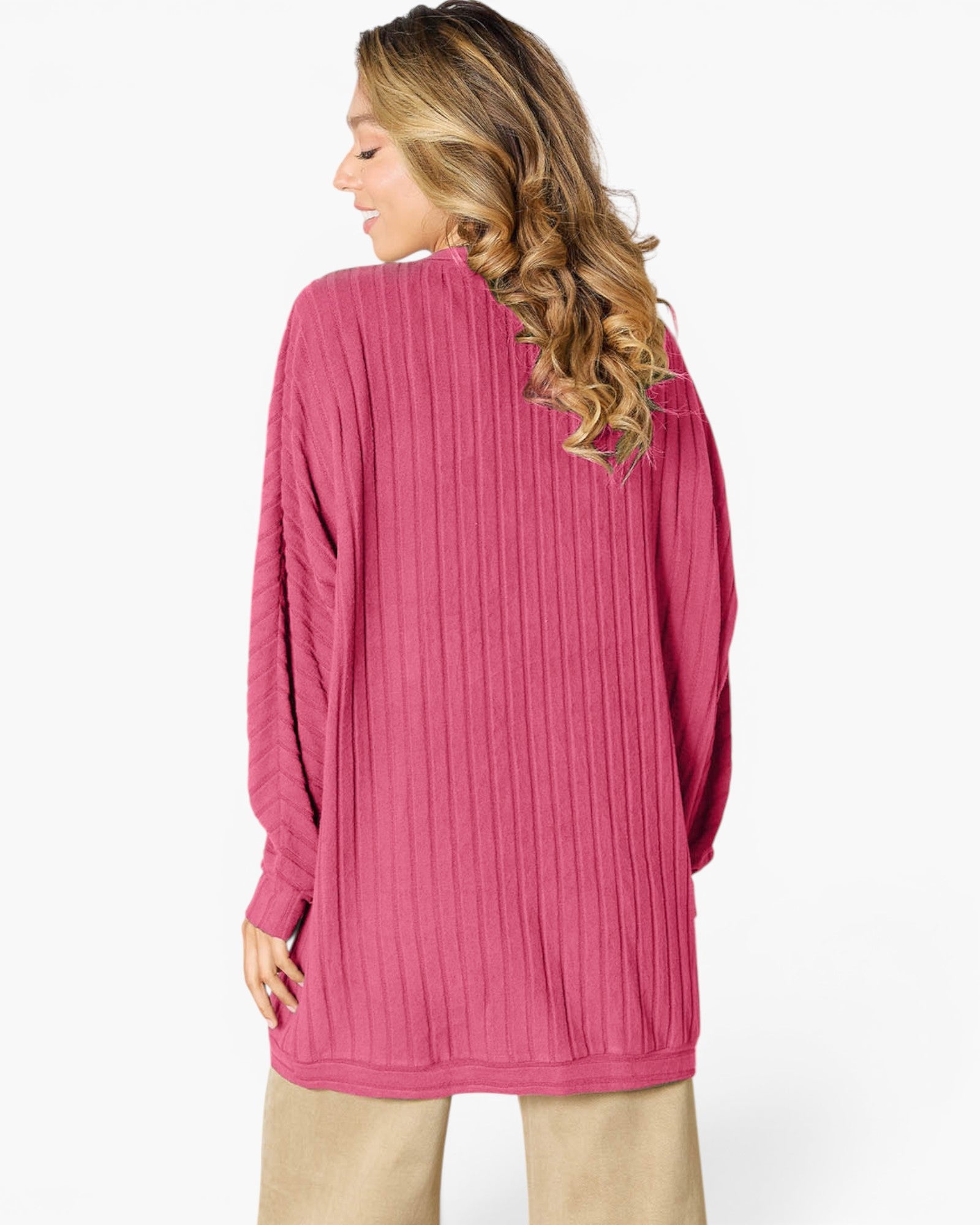 A woman shown from the back wearing a pink ribbed knit long-sleeve tunic sweater, paired with beige pants, against a plain white studio background.