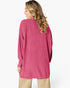 A woman shown from the back wearing a pink ribbed knit long-sleeve tunic sweater, paired with beige pants, against a plain white studio background.