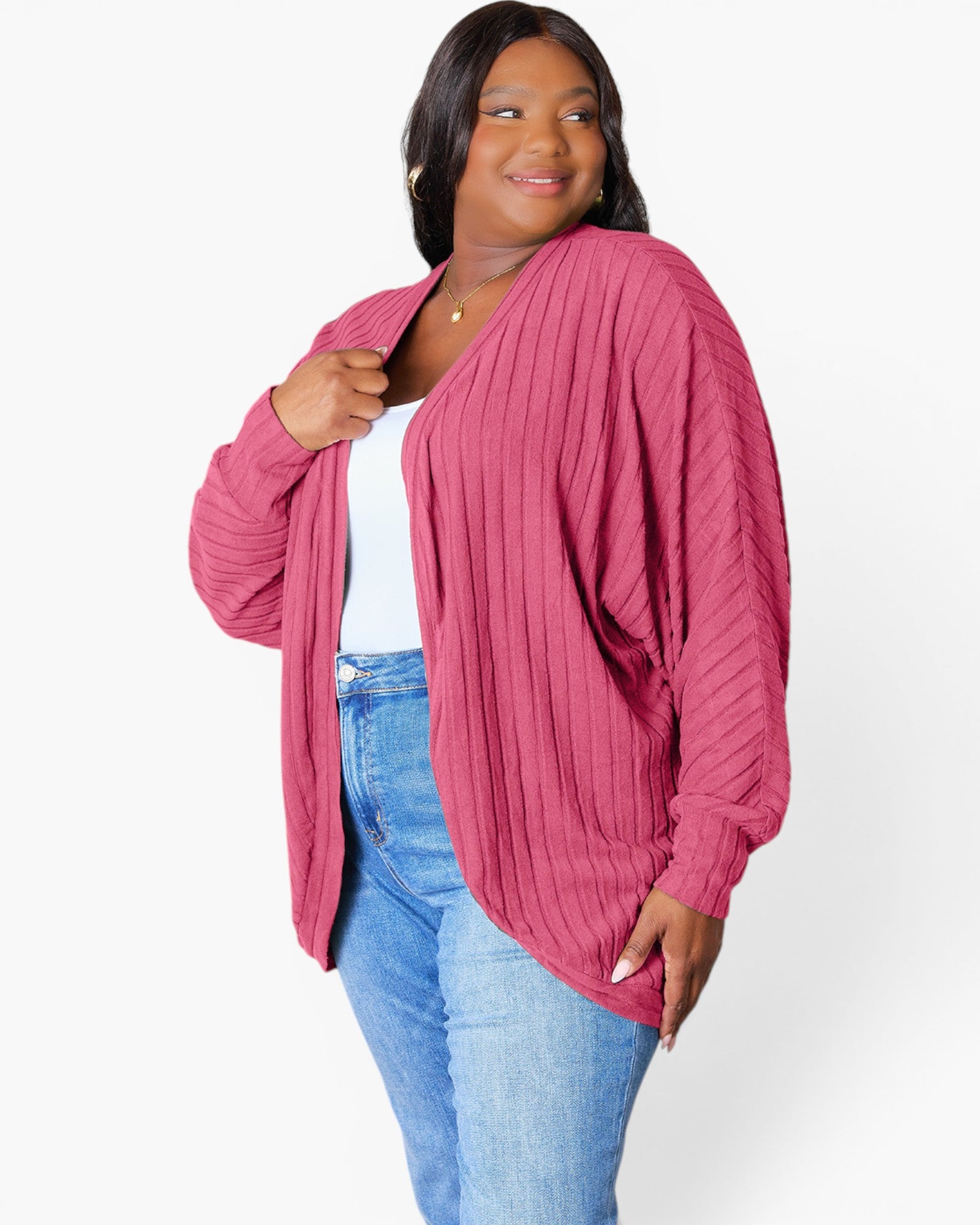 A woman wearing a pink ribbed knit cardigan over a white top and blue jeans, standing against a plain light gray studio background.