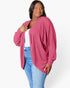 A woman wearing a pink ribbed knit cardigan over a white top and blue jeans, standing against a plain light gray studio background.