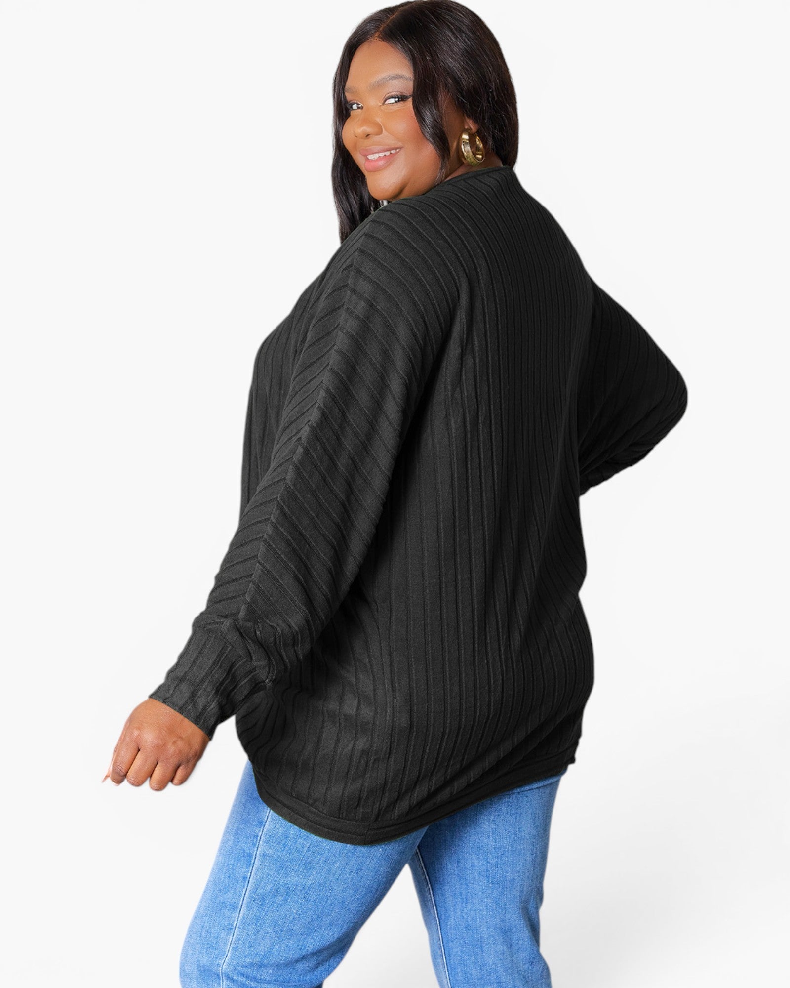 A woman wearing a black ribbed knit long-sleeve top and blue jeans, photographed against a plain light gray studio background.