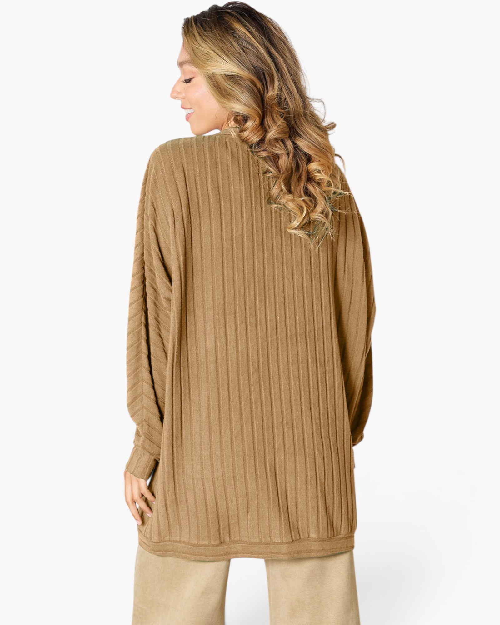 A woman shown from the back wearing a long-sleeve, oversized ribbed knit sweater in a warm camel brown color, paired with beige pants, against a plain white studio background.