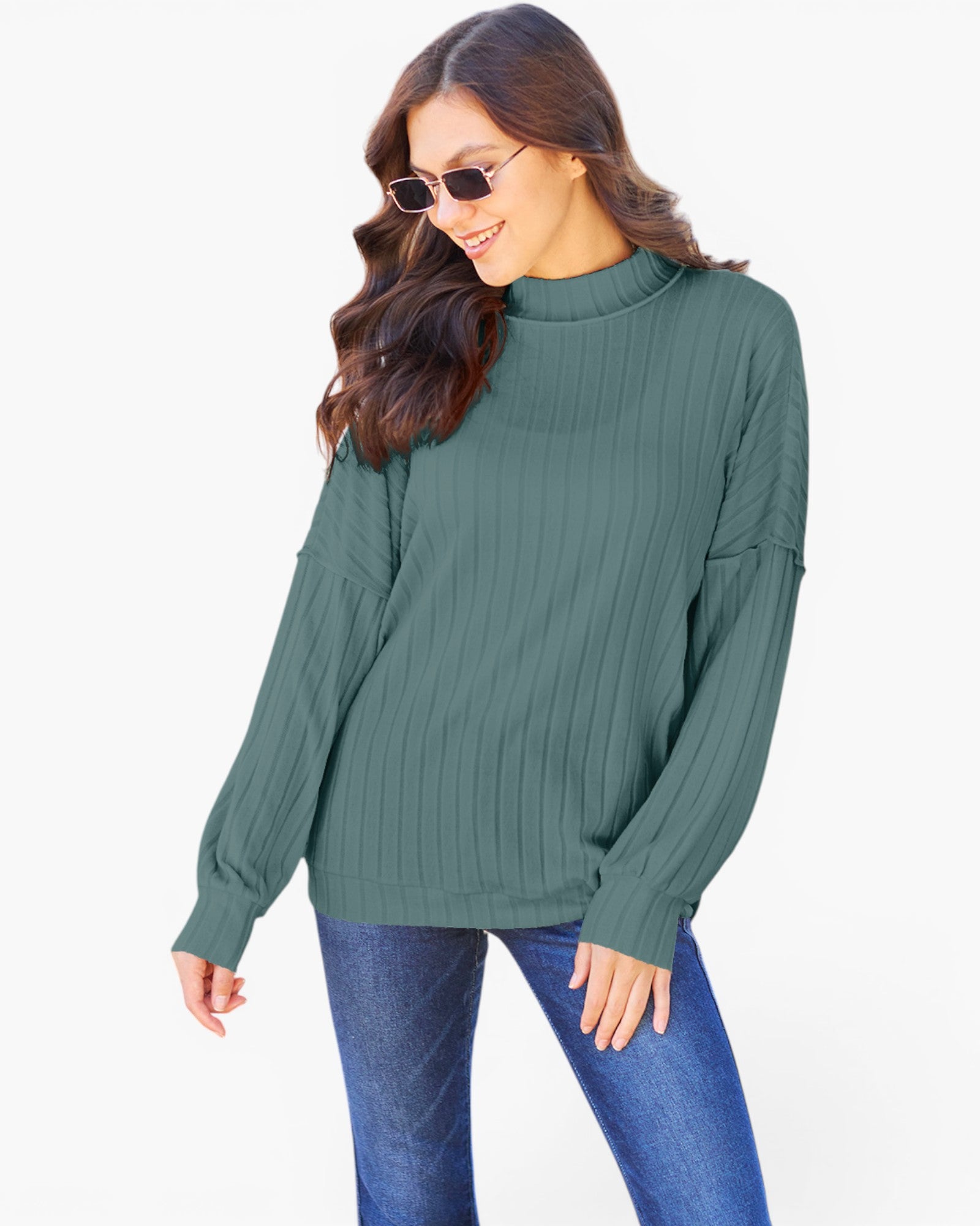 A woman wearing a ribbed long-sleeve mock neck sweater in sage green, paired with blue jeans, against a plain white background.