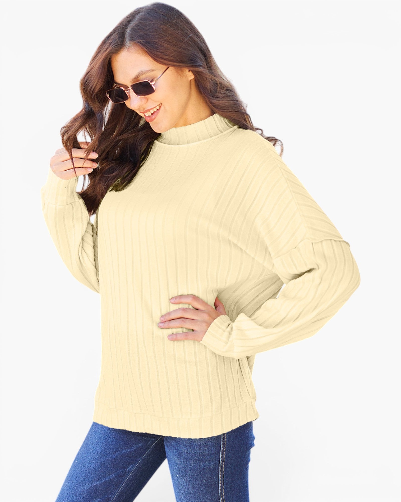 A woman wearing a pale yellow ribbed knit mock neck sweater and blue jeans, posing against a solid white background.