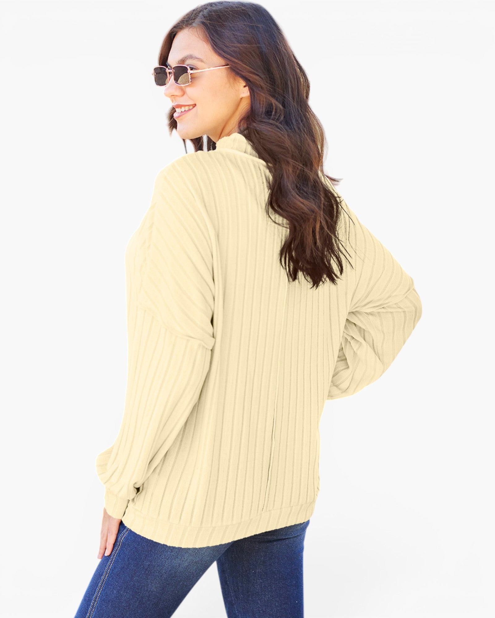 A woman wearing a cream-colored ribbed knit long-sleeve top and blue jeans, photographed from a side-back angle against a plain white background.