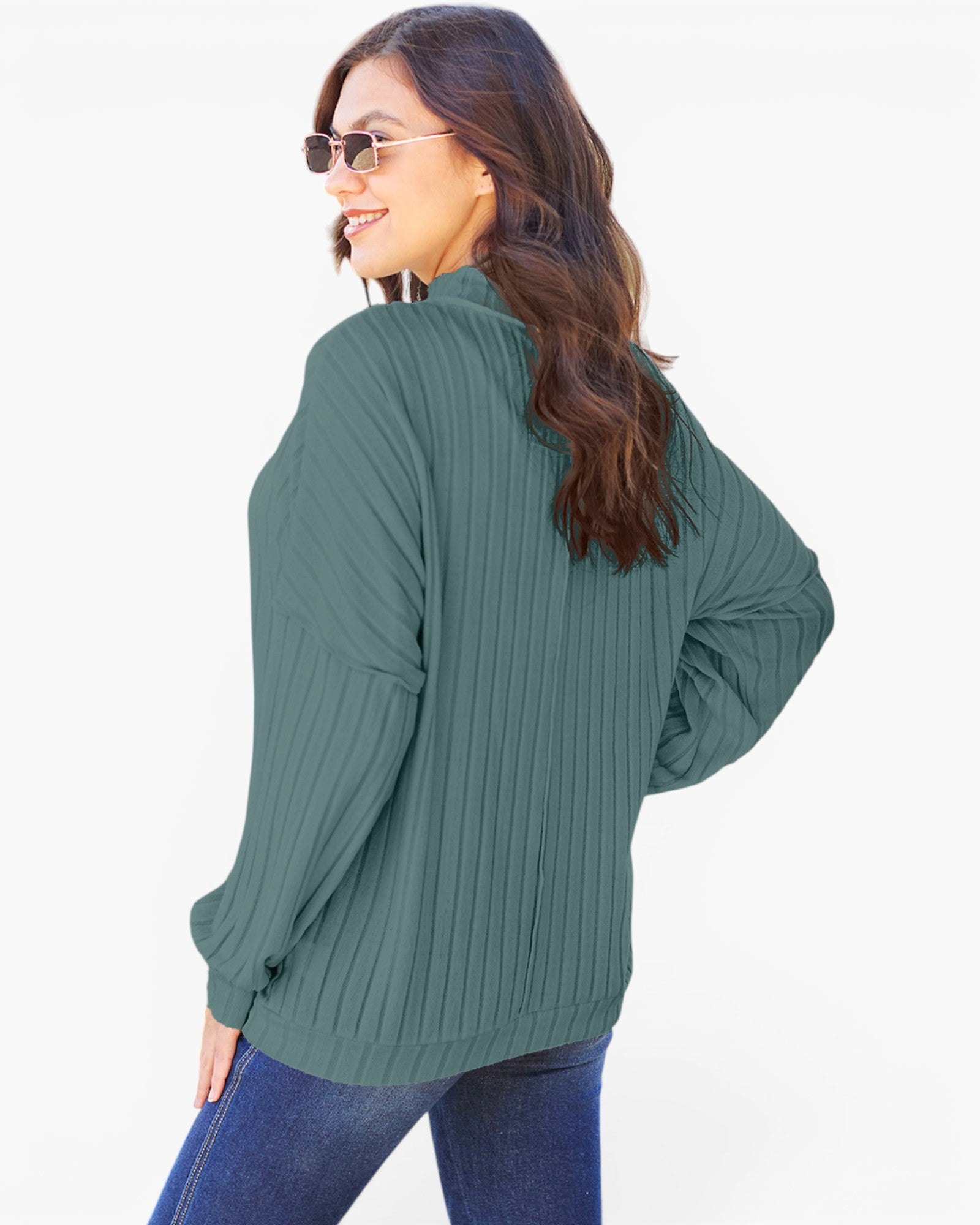 A woman wearing a sage green ribbed knit long-sleeve top and blue jeans, photographed from a three-quarters back angle against a plain white background.