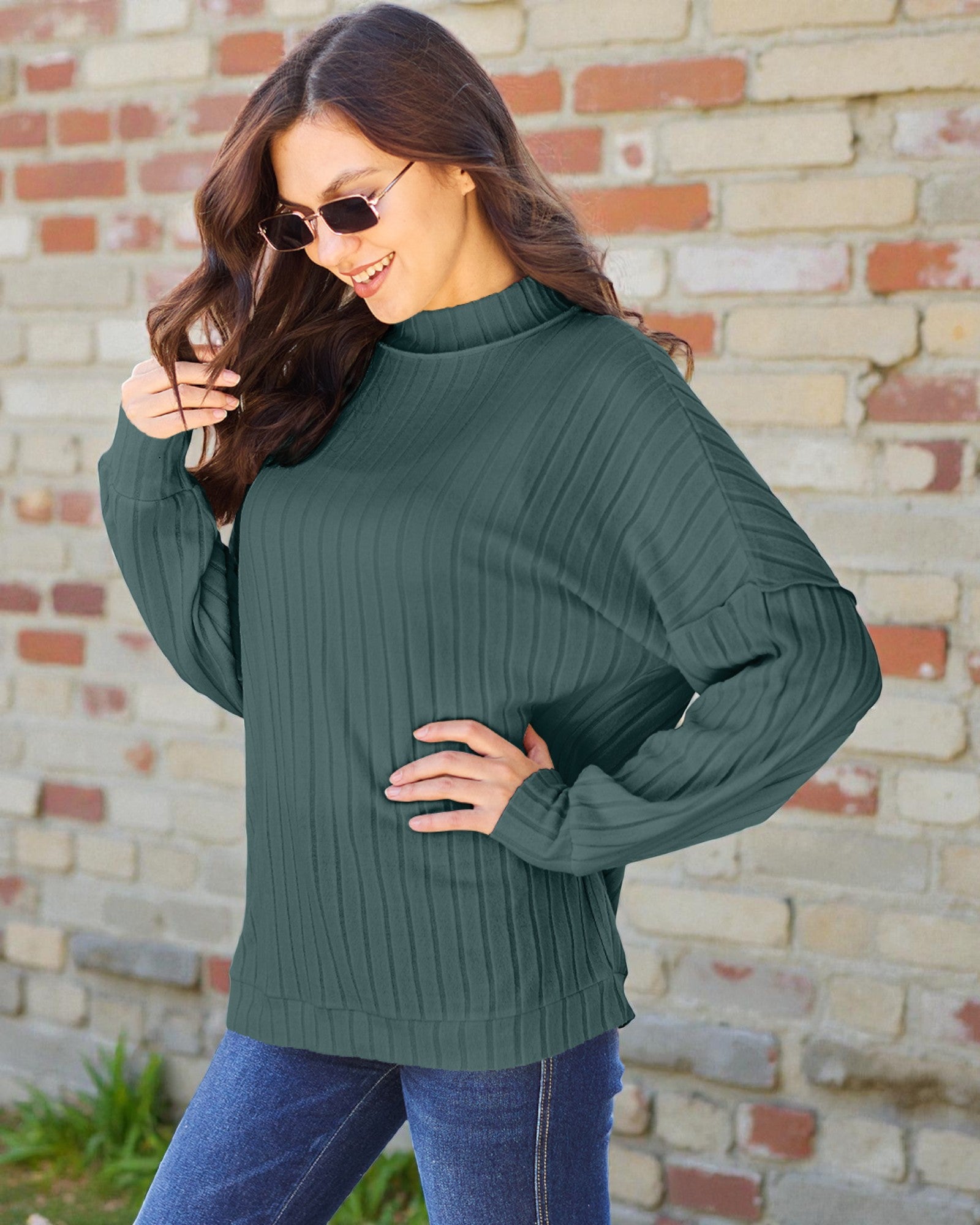 Woman wearing a green sweater and sunglasses against a brick wall.