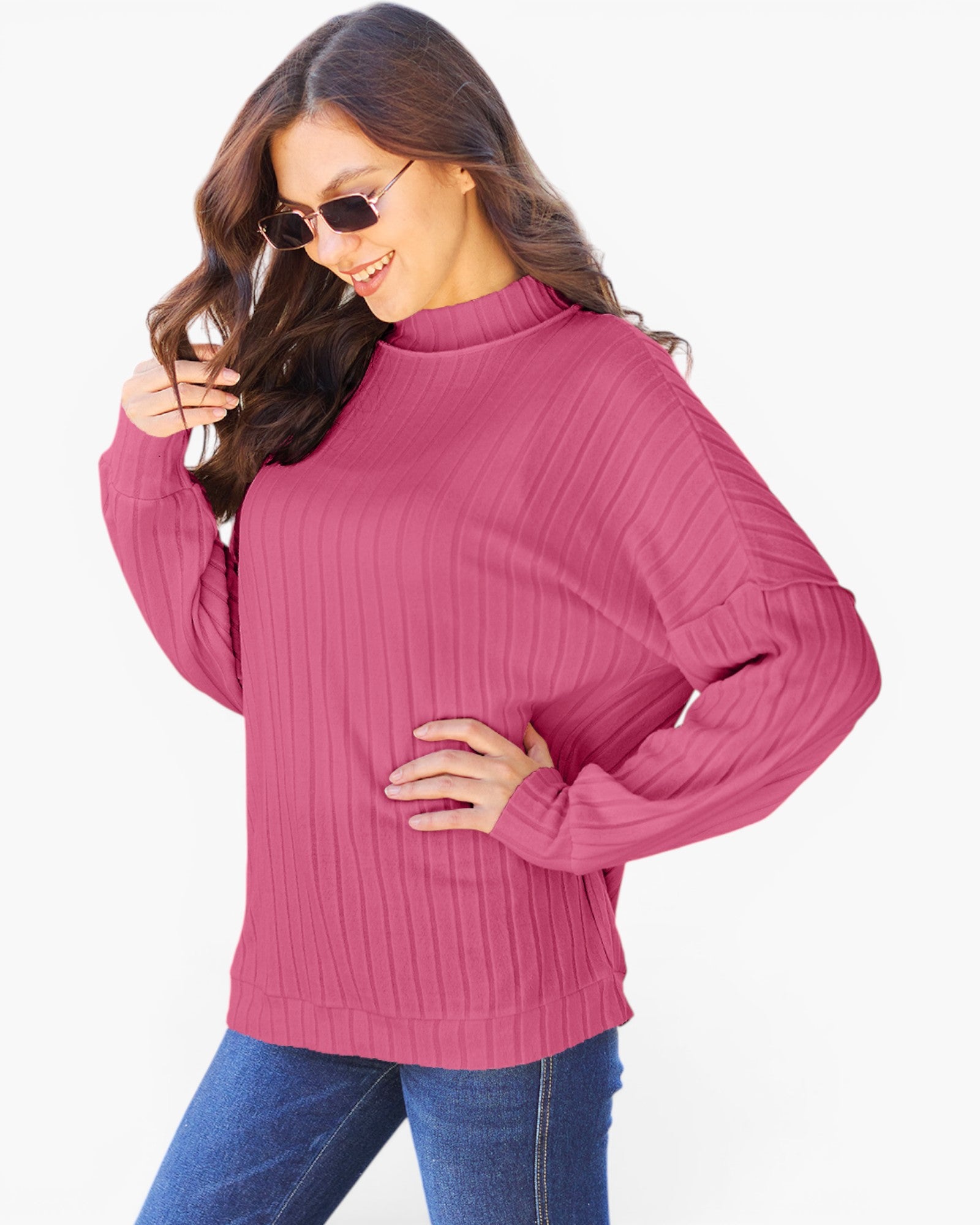 A woman wearing a pink ribbed knit mock neck sweater with long sleeves and blue jeans, posing against a light gray background.