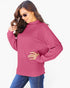 A woman wearing a pink ribbed knit mock neck sweater with long sleeves and blue jeans, posing against a light gray background.