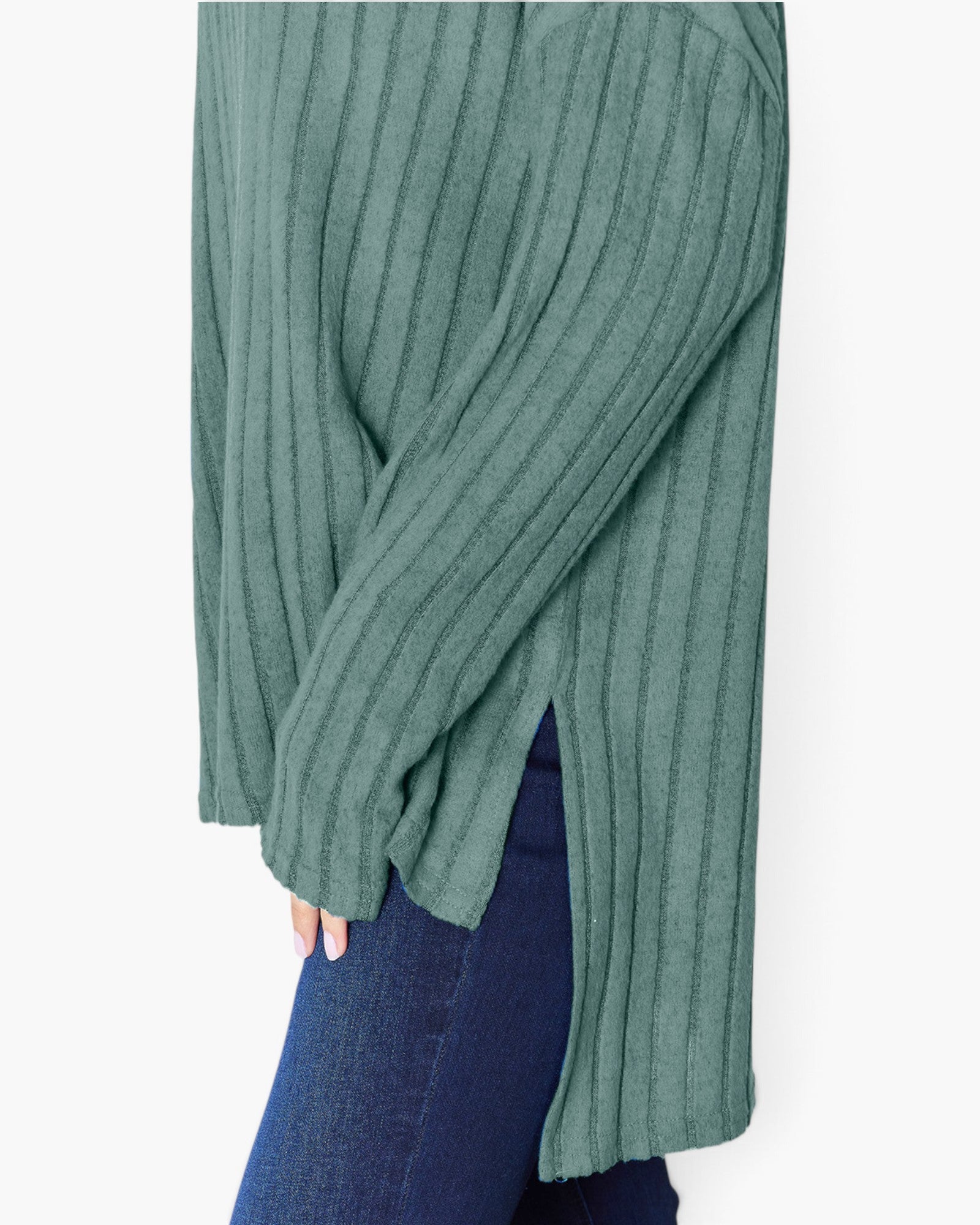 A close-up of a person wearing a ribbed knit sweater in a muted sage green color, paired with dark blue jeans, against a plain white background.