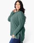A woman modeling a ribbed knit long-sleeve sweater in a muted sage green color, paired with blue jeans, against a plain white studio background.