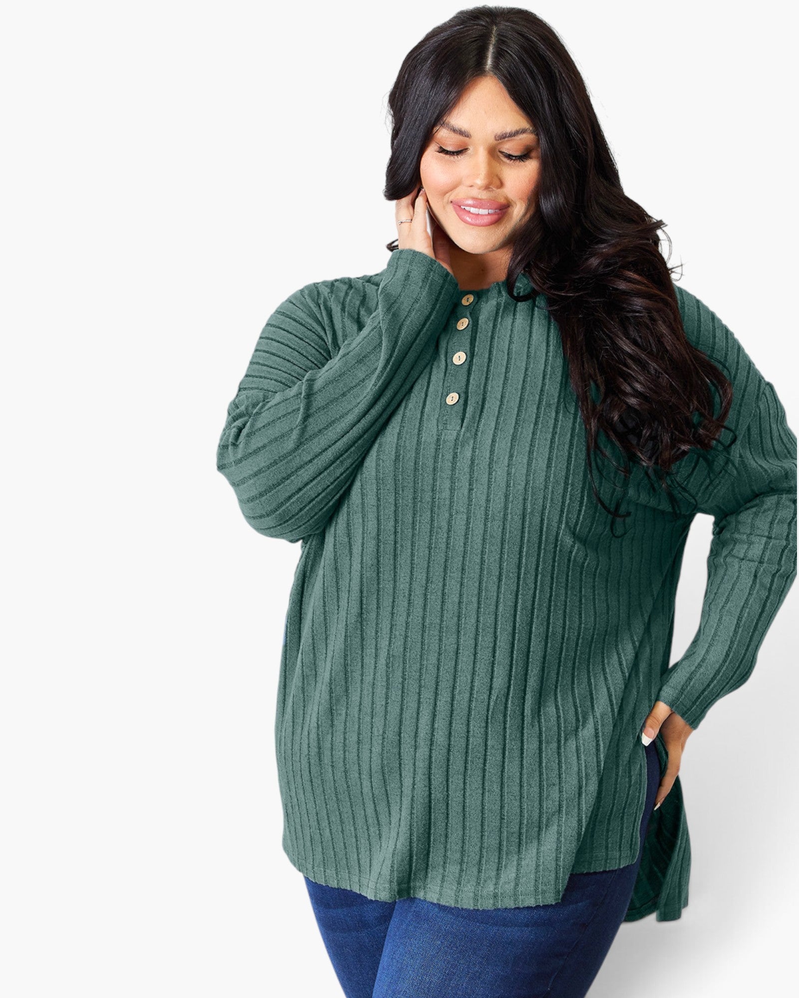 A woman wearing a ribbed knit long-sleeve henley top in sage green with gold buttons, paired with blue jeans, against a plain white studio background.