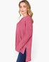 A woman wearing a pink ribbed knit long-sleeve top with side slits, paired with blue jeans, photographed against a plain white studio background.