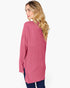 A woman with blonde wavy hair is shown from a side-back angle wearing a pink ribbed knit tunic sweater and dark blue jeans against a plain white background.