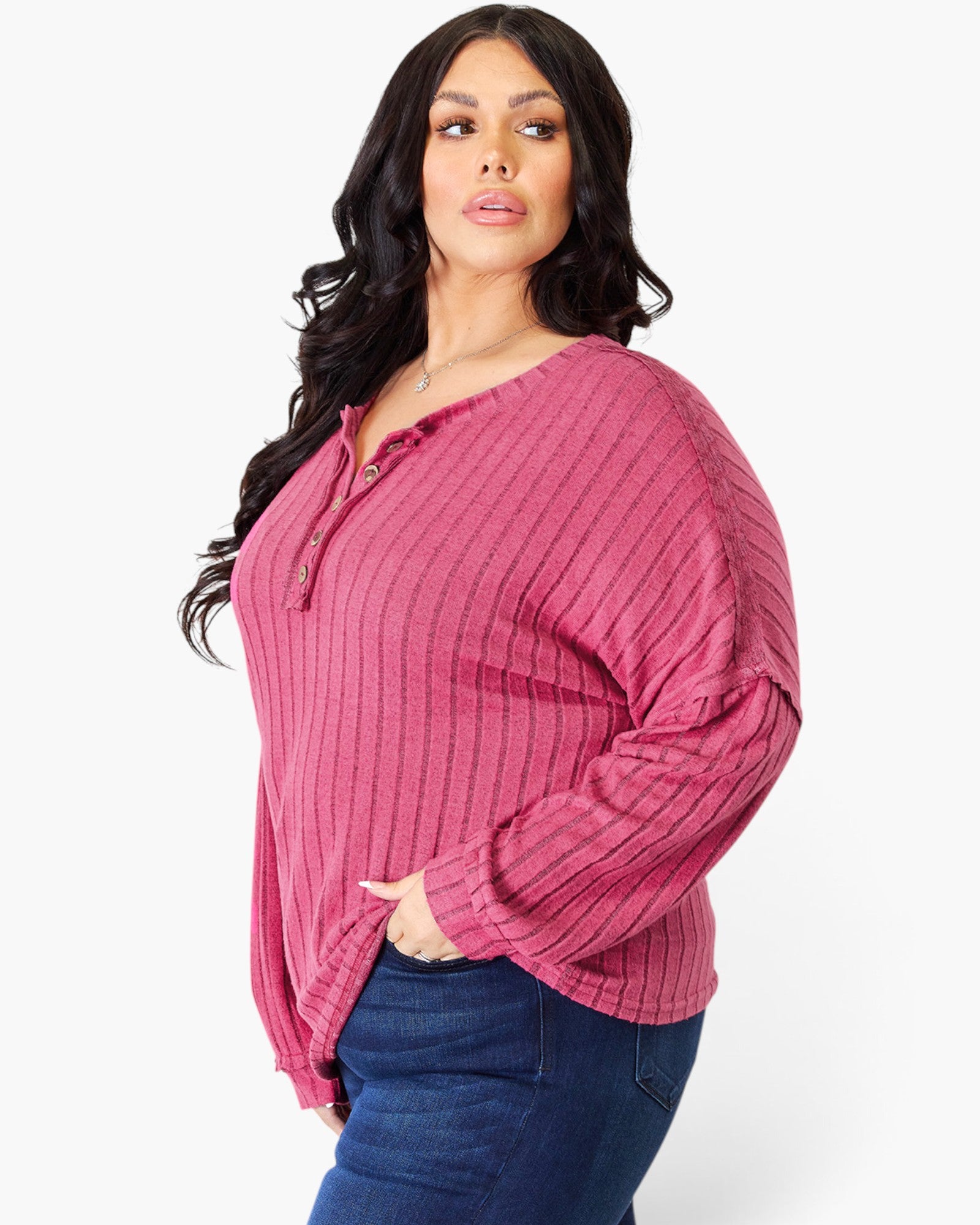 A woman modeling a ribbed long-sleeve henley top in dusty rose pink with blue jeans, photographed against a clean white studio background.