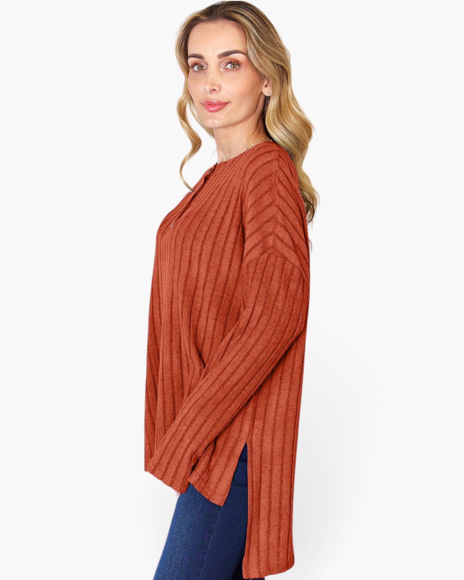 A woman wearing a rust-orange ribbed knit long-sleeve top with side slits, paired with blue jeans, photographed against a clean white studio background.