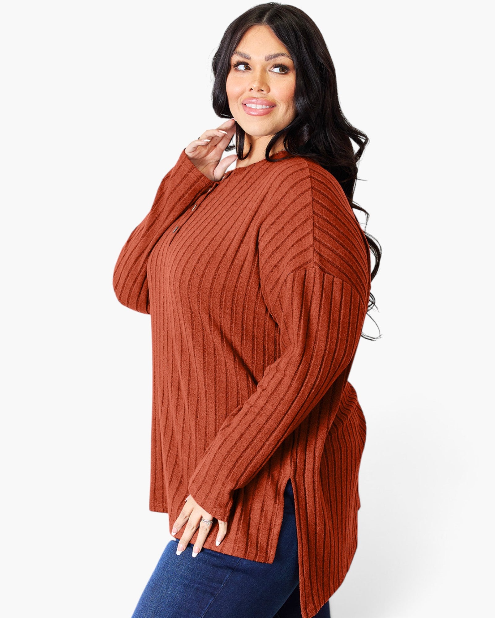 A woman modeling a rust-colored ribbed knit sweater with a crew neck and long sleeves, paired with blue jeans, against a plain white background.