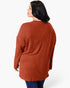 A woman shown from the back wearing a rust-orange ribbed knit sweater and dark blue jeans against a plain white studio background.
