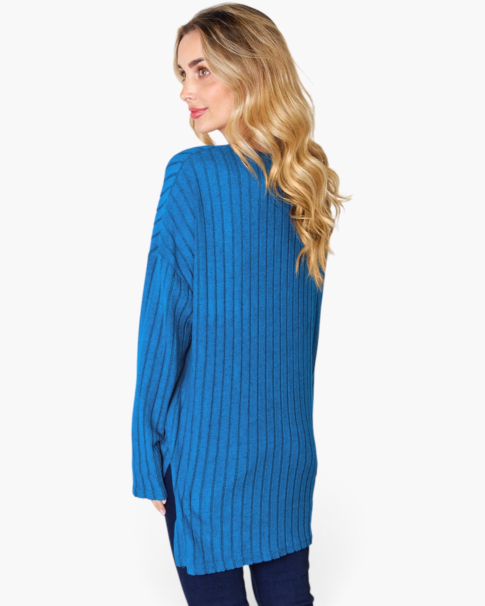 A woman with blonde wavy hair is shown from the back wearing a bright blue ribbed knit tunic sweater and dark blue jeans against a plain white background.