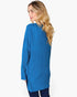A woman with blonde wavy hair is shown from the back wearing a bright blue ribbed knit tunic sweater and dark blue jeans against a plain white background.