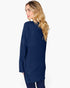 A woman with long blonde wavy hair is shown from a side-back angle wearing a navy blue ribbed knit sweater and dark pants against a plain white studio background.