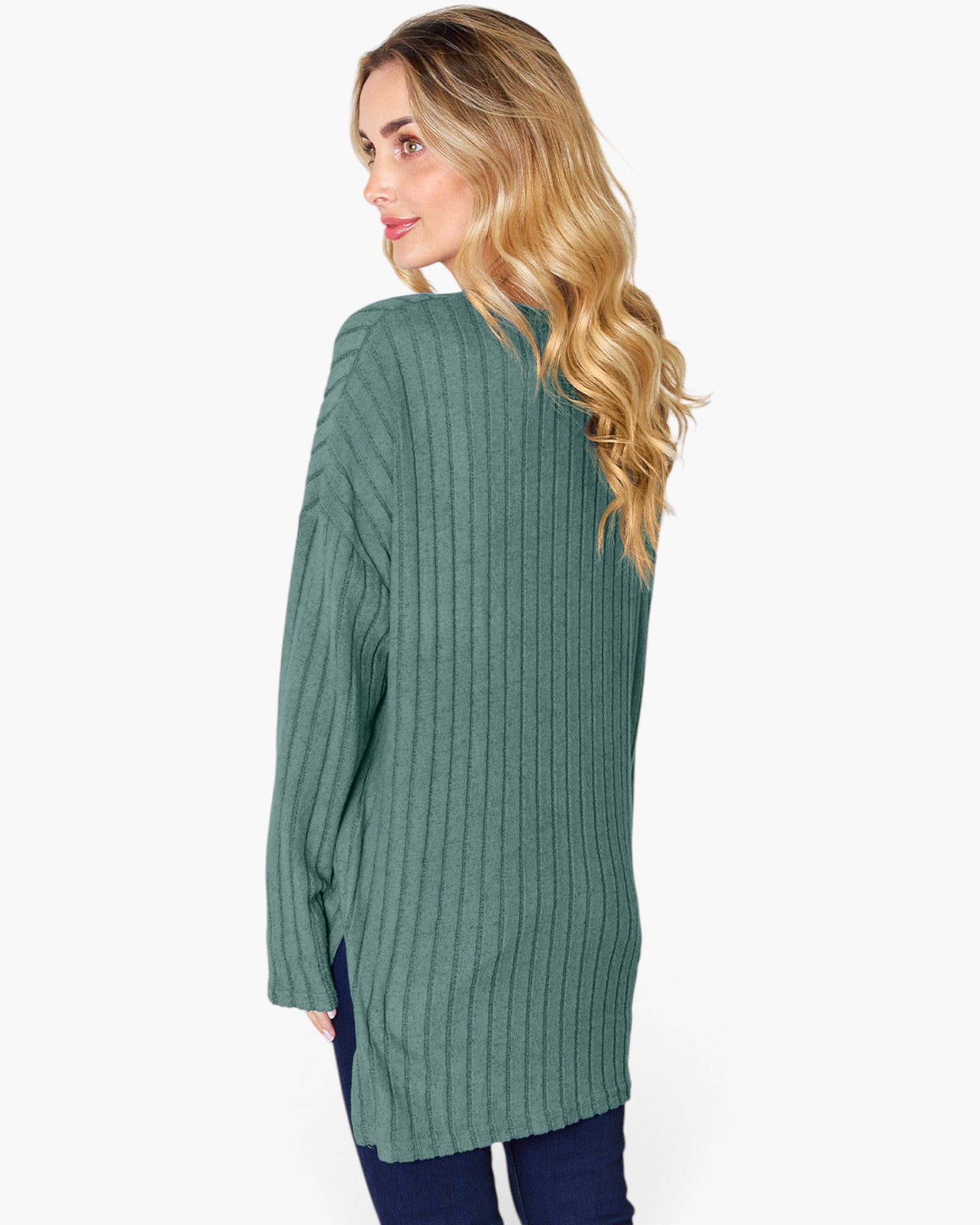 A woman with blonde wavy hair is shown from a back-three-quarter view wearing a sage green ribbed knit sweater and dark blue jeans against a plain white background.