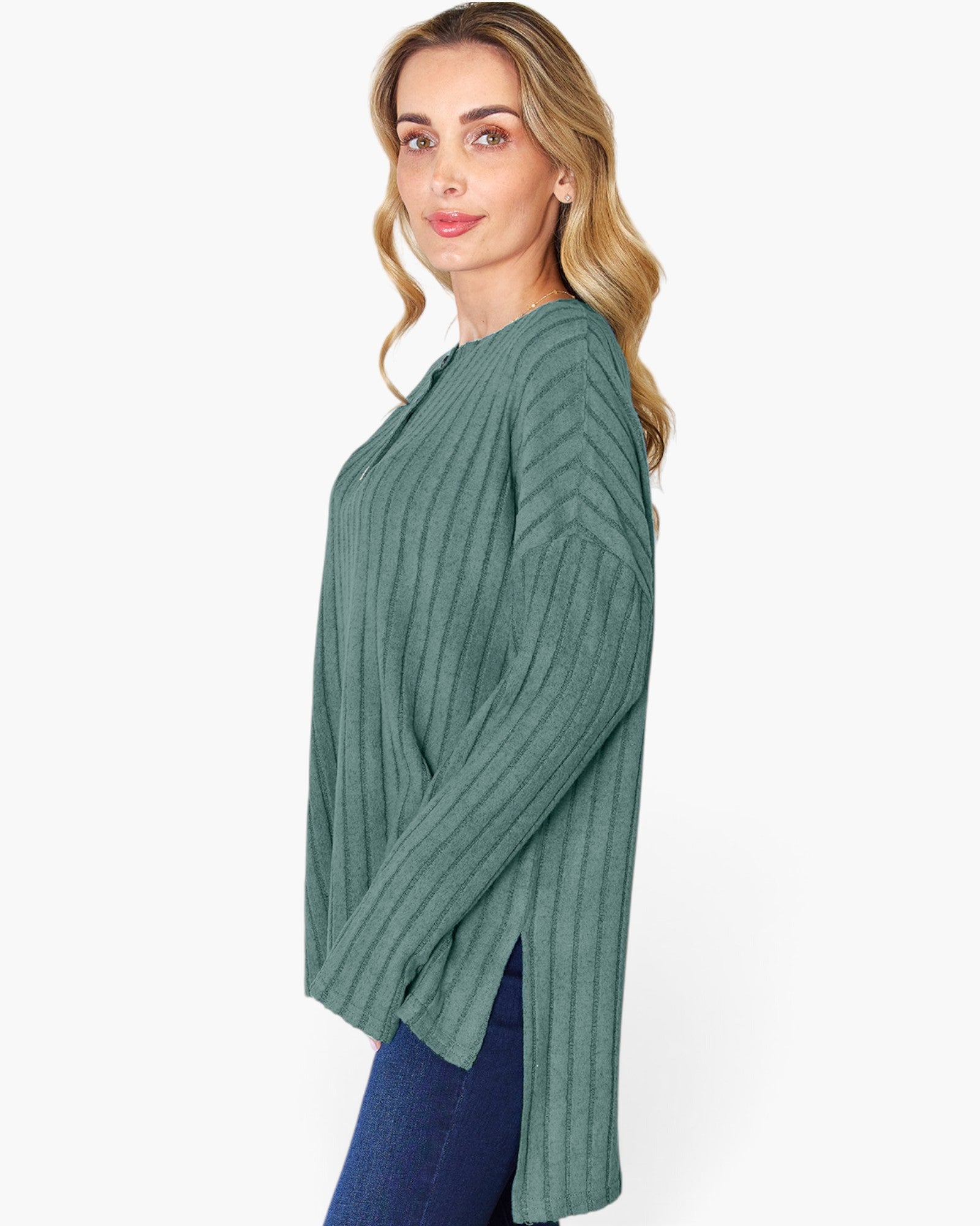 A woman wearing a sage green ribbed knit long-sleeve top with side slits, paired with blue jeans, photographed against a plain white studio background.