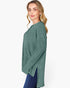 A woman wearing a sage green ribbed knit long-sleeve top with side slits, paired with blue jeans, photographed against a plain white studio background.