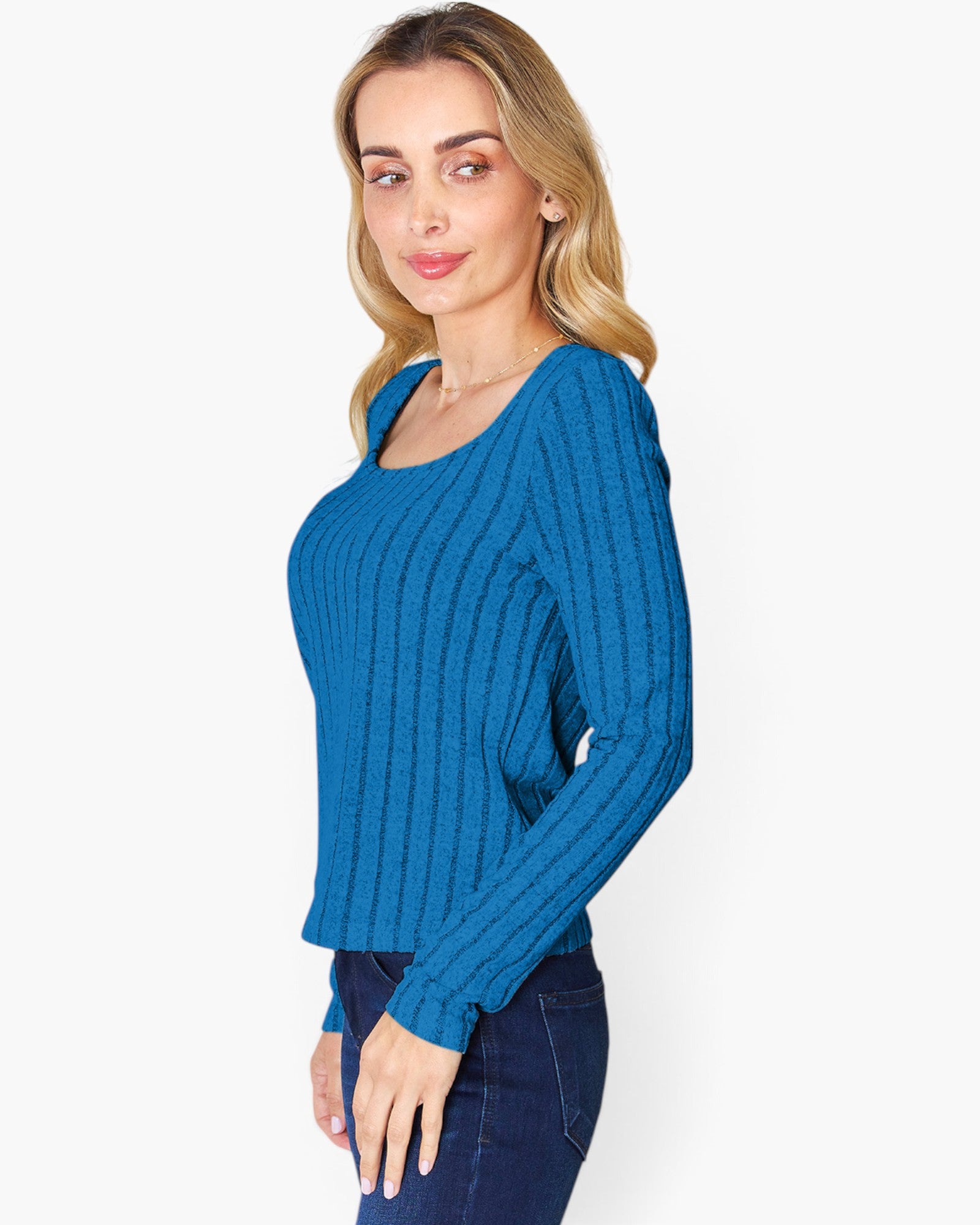 A woman wearing a bright blue ribbed knit long-sleeve top and dark blue jeans, photographed against a plain light gray studio background.