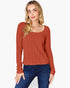 A woman modeling a rust-orange ribbed knit long-sleeve sweater with a scoop neckline, paired with dark blue jeans, against a plain white background.