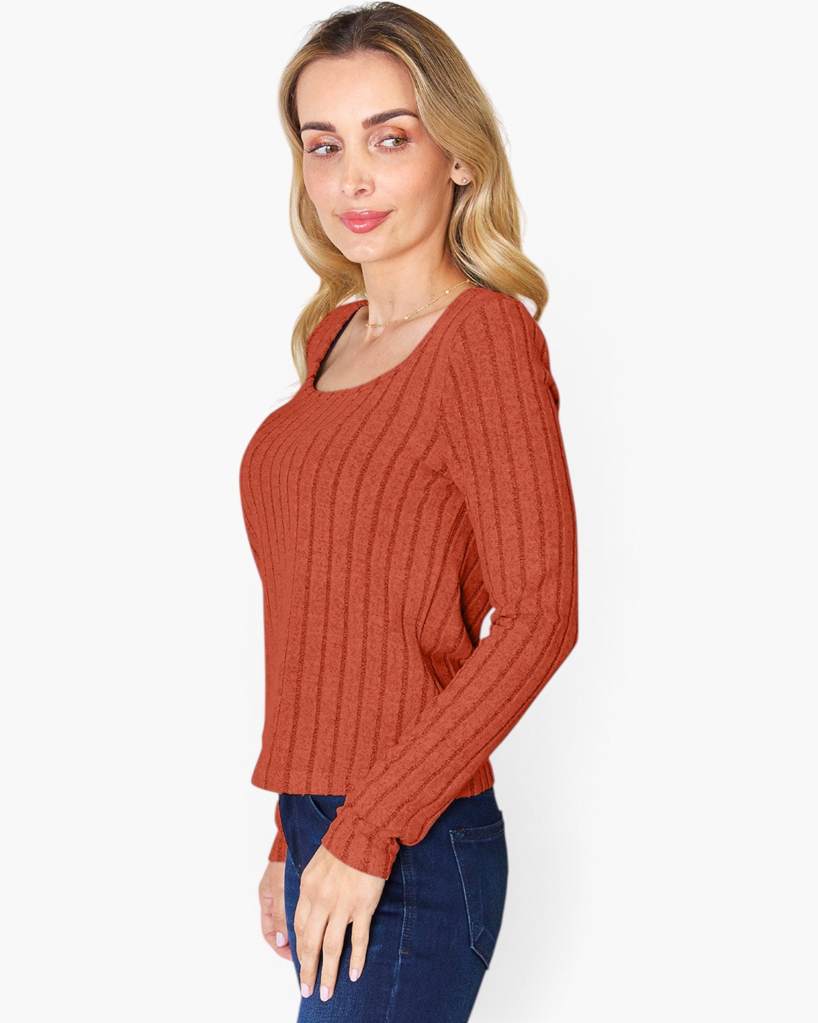 A woman modeling a rust-orange ribbed knit long-sleeve top with a scoop neckline, paired with dark blue jeans, against a plain light gray background.