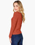 A woman with blonde hair is shown from a side-back angle wearing a rust-orange ribbed knit sweater and dark blue jeans against a plain white background.