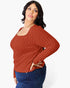 A woman modeling a rust-orange ribbed knit long-sleeve top with a scoop neckline, paired with dark blue jeans, against a plain white studio background.