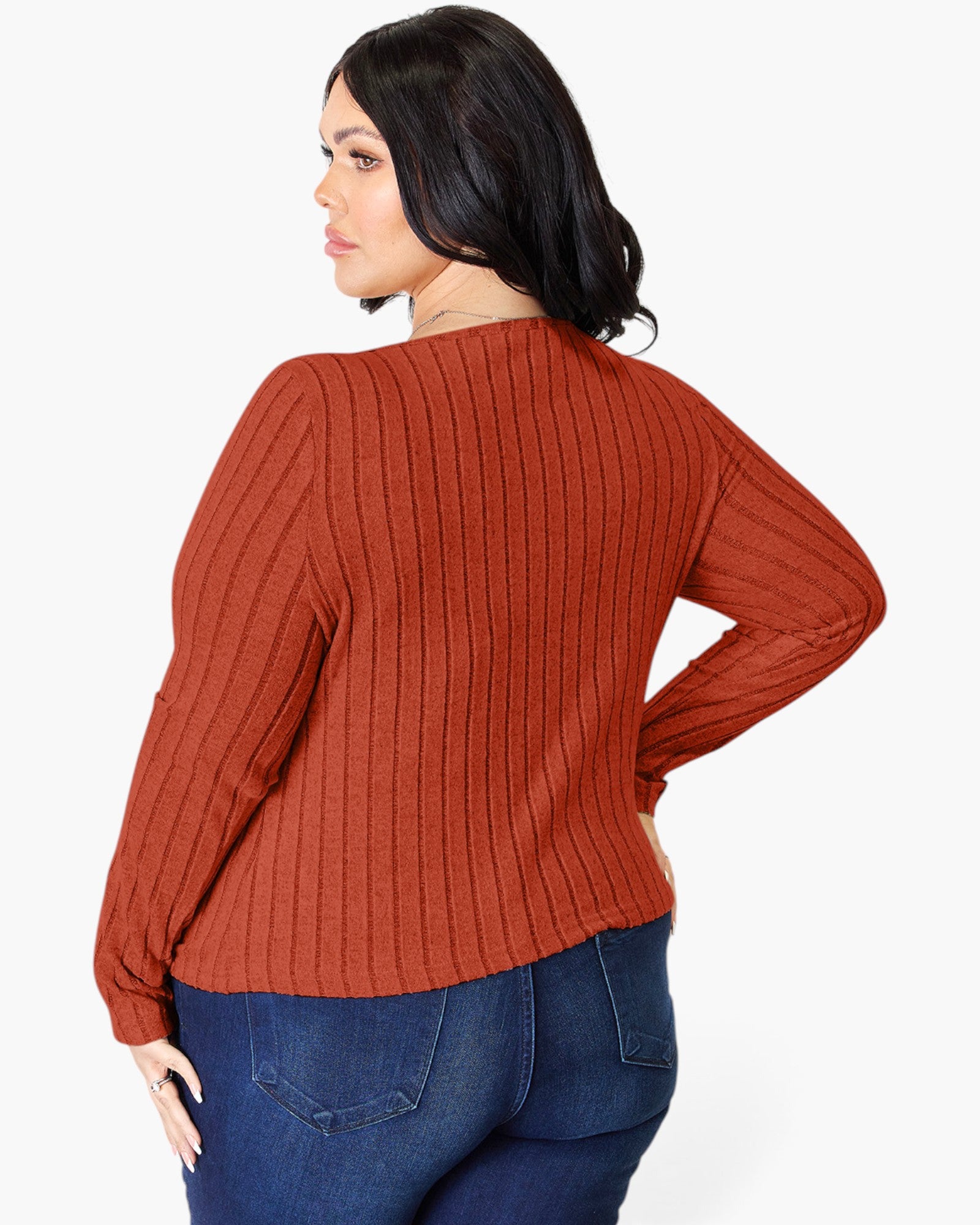 A woman wearing a rust-orange ribbed knit sweater and blue jeans, shown from a back-three-quarter angle against a plain white studio background.