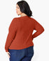 A woman wearing a rust-orange ribbed knit sweater and blue jeans, shown from a back-three-quarter angle against a plain white studio background.