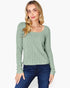 A woman wearing a sage green ribbed knit long-sleeve top with a square neckline, paired with dark blue jeans, against a plain white background.
