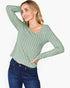 A woman wearing a sage green ribbed knit long-sleeve top and dark blue jeans, posing against a light gray background.
