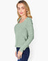 A woman wearing a sage green ribbed knit long-sleeve sweater with a scoop neckline, paired with dark blue jeans, photographed against a solid light gray background.