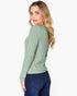 A woman with blonde hair is shown from a side-back angle wearing a sage green ribbed knit sweater and dark blue jeans against a plain white background.