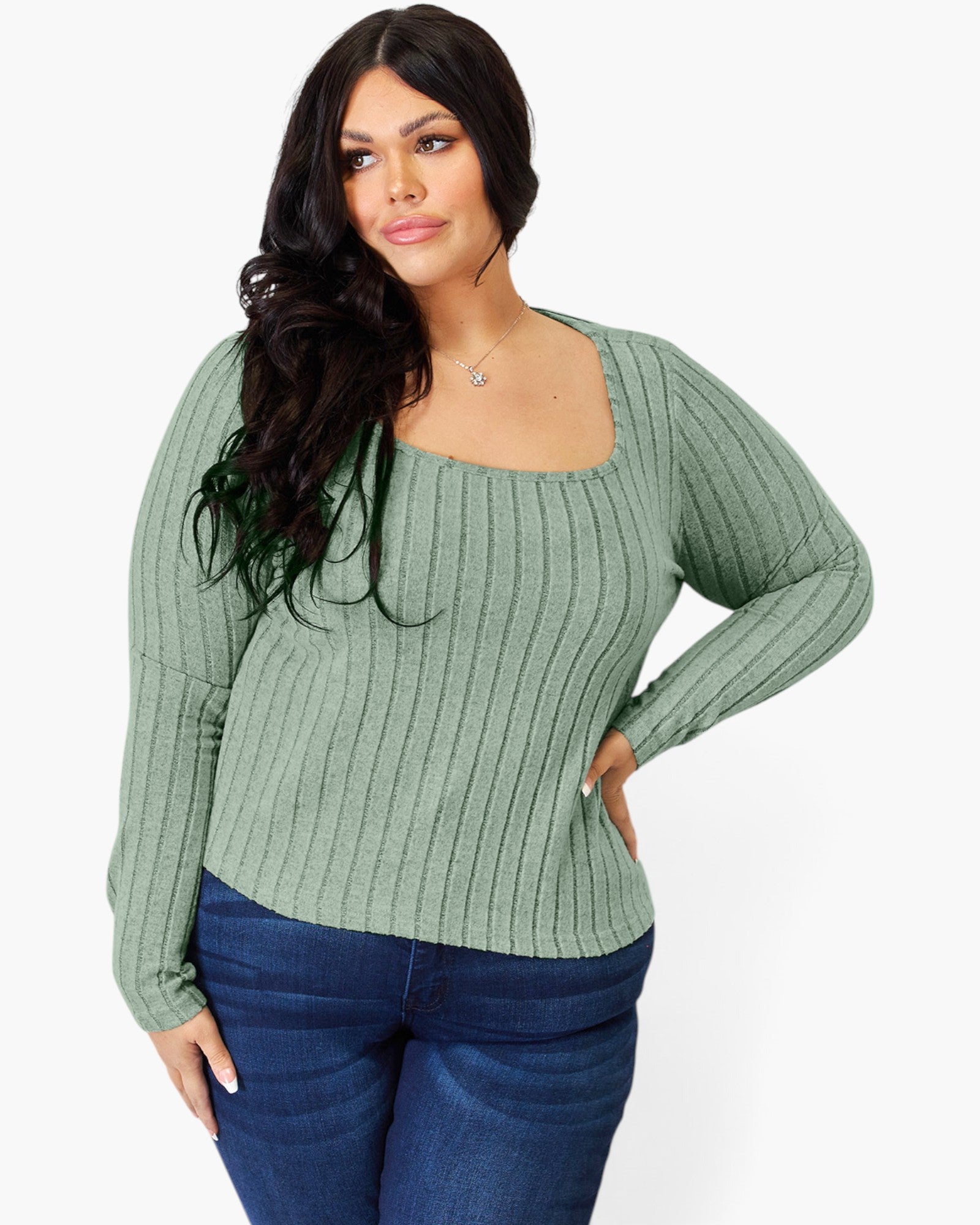 A woman modeling a sage green ribbed knit long-sleeve top with a square neckline, paired with dark blue jeans, against a plain white background.