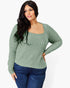 A woman modeling a sage green ribbed knit long-sleeve top with a square neckline, paired with dark blue jeans, against a plain white background.