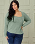 Woman wearing a green ribbed sweater and blue jeans against a light-colored wall.