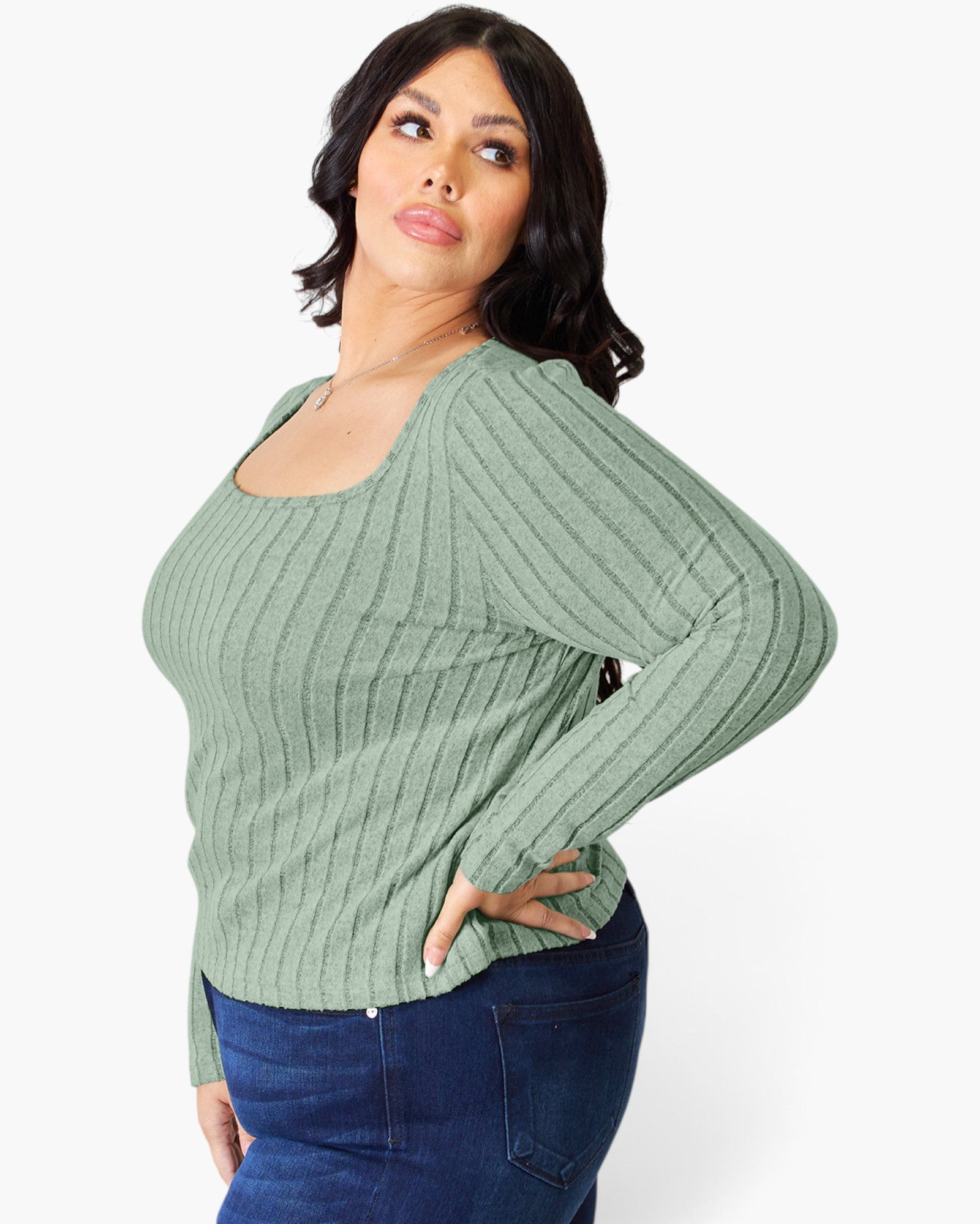 A woman modeling a sage green ribbed knit long-sleeve top with a scoop neckline, paired with dark blue jeans, against a plain white studio background.