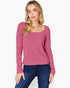 A woman modeling a pink ribbed knit long-sleeve top with a scoop neckline, paired with dark blue jeans, against a plain light gray background.