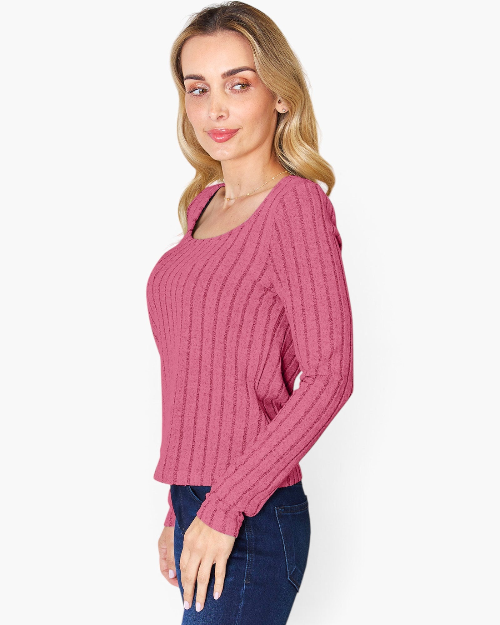 A woman modeling a pink ribbed knit long-sleeve top with a scoop neckline, paired with dark blue jeans, against a plain light gray background.